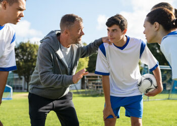 Mature coach teaching strategy to high school team on football field. Young soccer players standing together united and listening coach motivational speech. Coach giving team advise before school match.